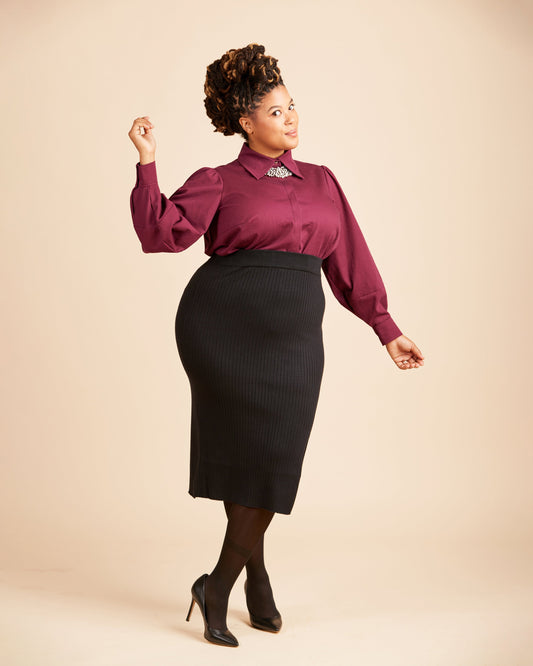 Woman wearing a burgundy blouse and black skirt on a beige background. This top is the Vernon Shirt sewing pattern from Cashmerette.