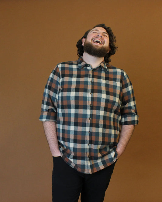 A man with a beard and curly hair laughing heartily, wearing a brown, black, and beige plaid button-down shirt with black pants, standing against a solid brown studio background.