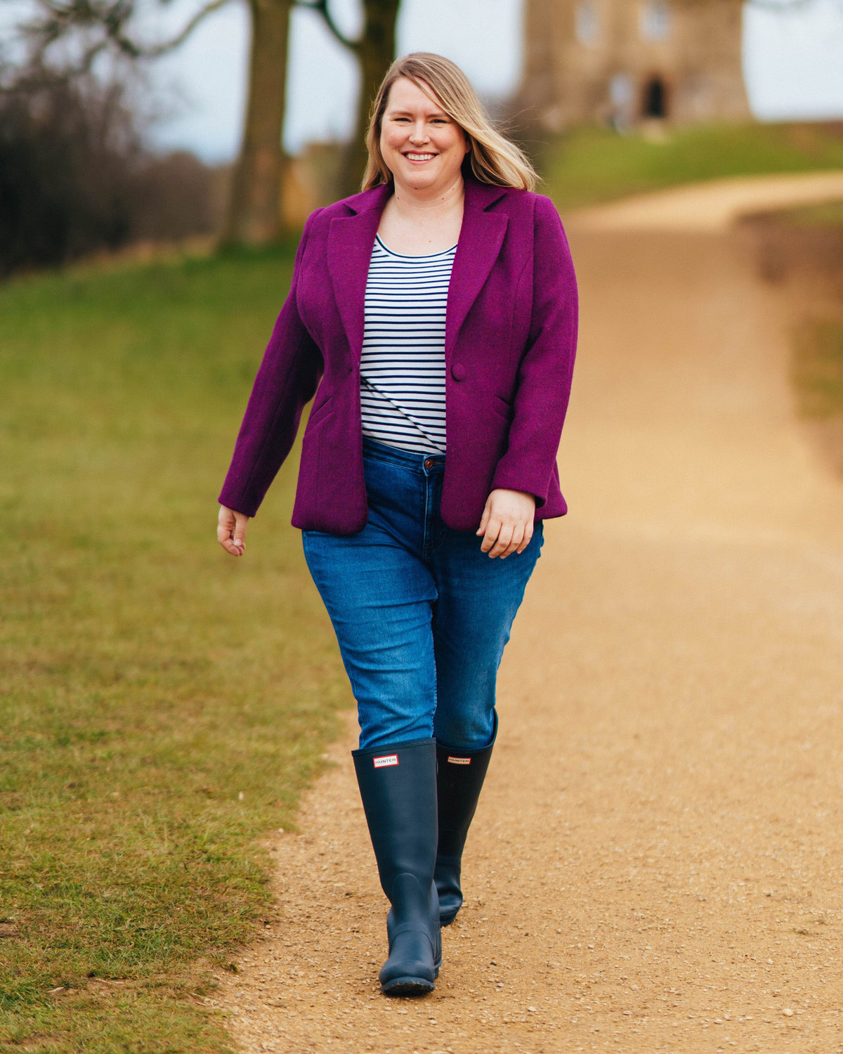 Woman in a purple coat, striped shirt, and blue jeans walking on a path outdoors.. This blazer is the Auburn Blazer sewing pattern from Cashmerette.
