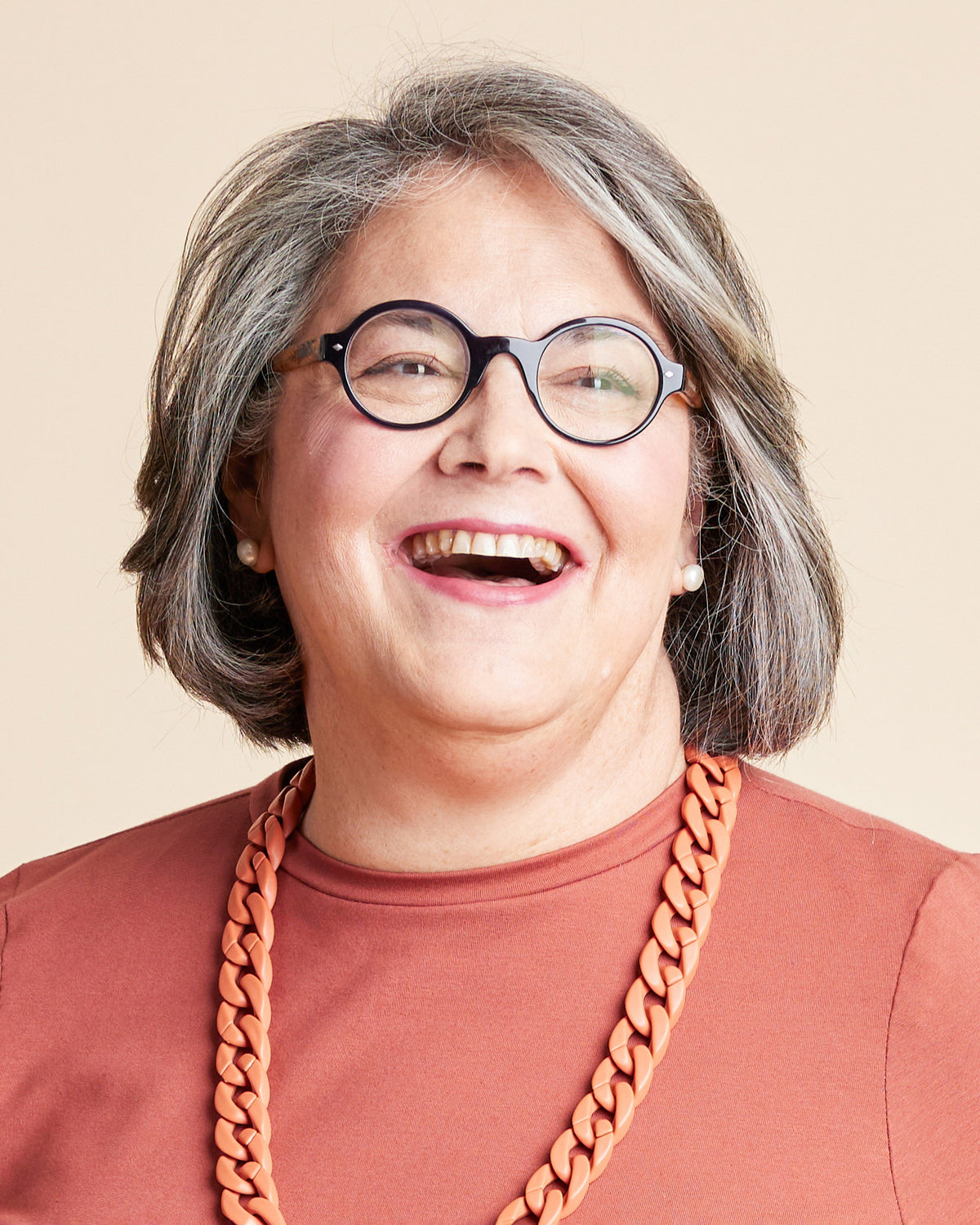Person with gray hair wearing glasses and a brown shirt with a large orange necklace on a beige background. This is the Grafton Dress, Top & Skirt sewing pattern from Cashmerette.