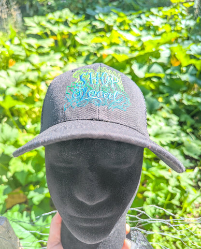 A black baseball cap with a colorful embroidered 'SHOP Local' logo, displayed on a black mannequin head outdoors with a green garden background.