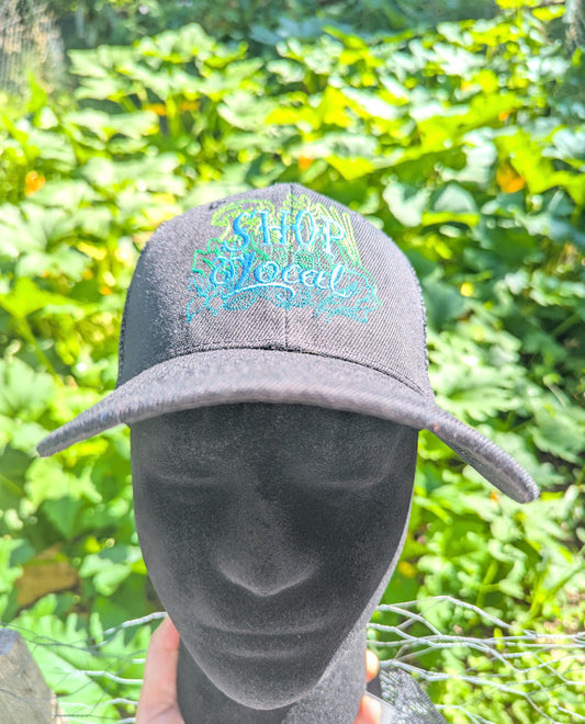 A black baseball cap with a colorful embroidered 'SHOP Local' logo, displayed on a black mannequin head outdoors with a green garden background.