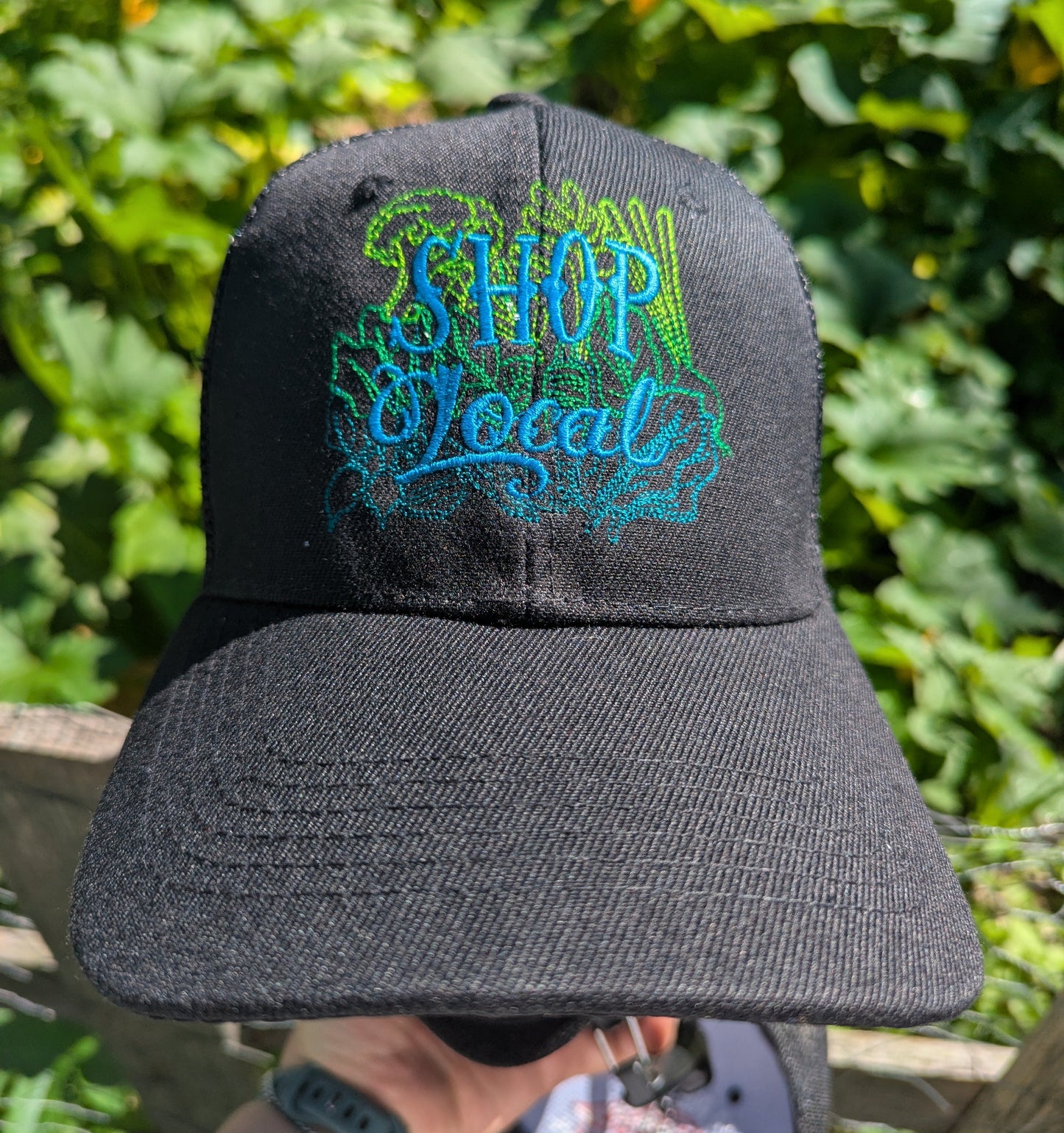 A black baseball cap with 'SHOP Local' embroidered on the front in blue and green, held up outdoors in front of a leafy green garden background.