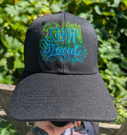 A black baseball cap with 'SHOP Local' embroidered on the front in blue and green, held up outdoors in front of a leafy green garden background.
